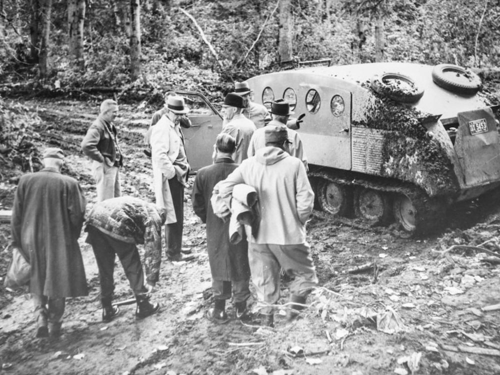 A black and white photo shows a cluster of men, most of them in business attire, inspecting a patch of cleared ground amidst foliage, with a tractor-like vehicle nearby.