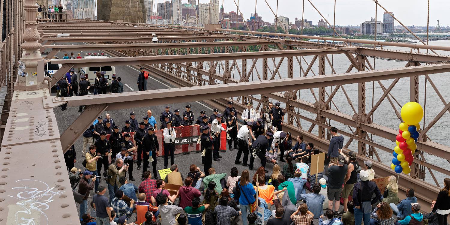 Shot from over a trellis on the Brooklyn Bridge, we look down on a skirmish between New York City police officers in dark uniforms and Occupy Wall Street demonstrators in street clothes in the foreground. Some officers are holding a sign that reads &ldquo;Police line do not cross.&rdquo; Other officers are lifting someone up with their arms and legs while demonstrators film the scene and flip the officers the bird.