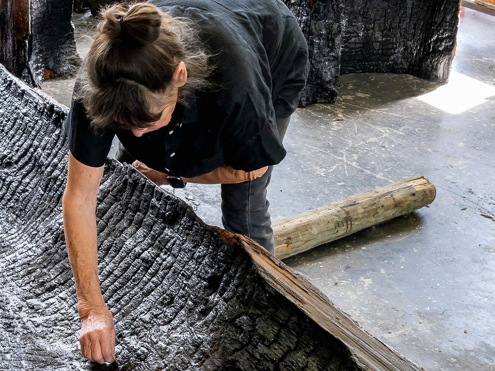 Martha Sturdy is bending and reaching into the hollowed-out interior of a large tree trunk laying across a studio floor. The trunk’s interior is black and burnt. Sturdy’s brown hair is tied up in a loose bun. She is wearing a black short-sleeved button-up shirt. We can’t see her face because she is bending toward the camera. Behind her are additional burnt tree trunks standing vertically on the grey floor.