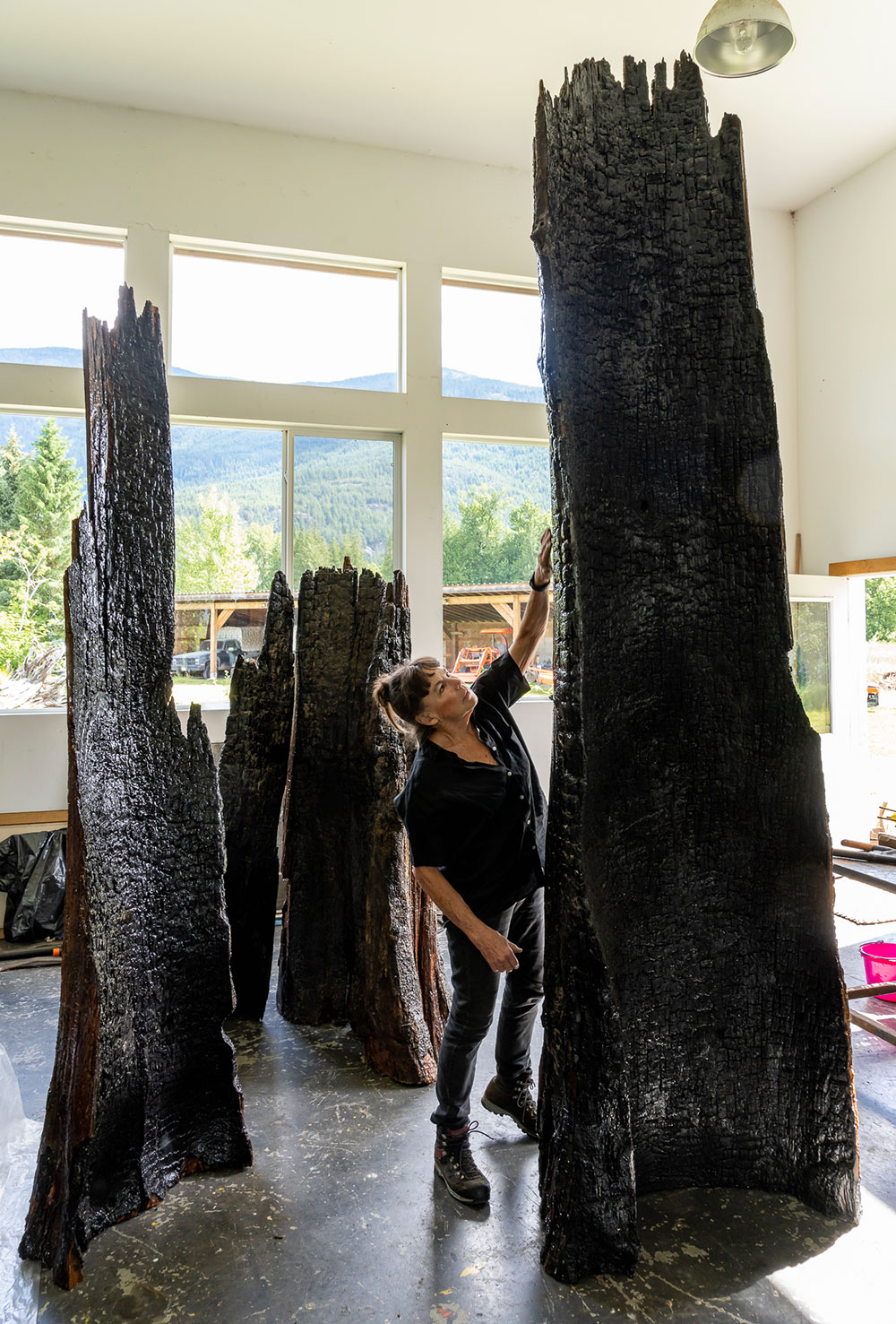 Martha Sturdy is in the centre of the frame, working in her studio earlier this year. Her brown hair is tied back in a loose bun and bangs frame her face. She is wearing black and reaching up to a charred piece of timbre part of her <em>All Fall Down</em> exhibition. She is surrounded by other similar pieces of wood. The studio floor is grey laminate. Behind her through the large windows cut into white walls, one can see a sunny mountain range.