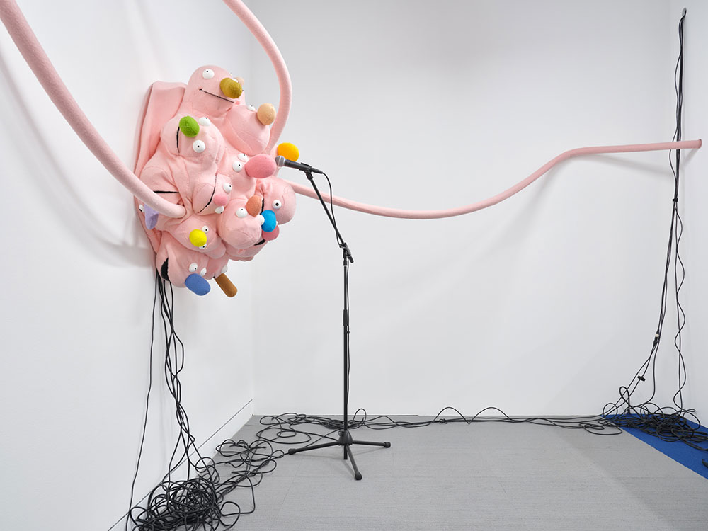 To the left of the frame, a baby-pink assembly of what appears to be children&rsquo;s puppet heads with colourful soft fabric noses are arranged in a round cluster near a microphone. Black wires dangle from the puppet heads down a white wall onto a gray floor. Long baby-pink coils of fabric are in the foreground.
