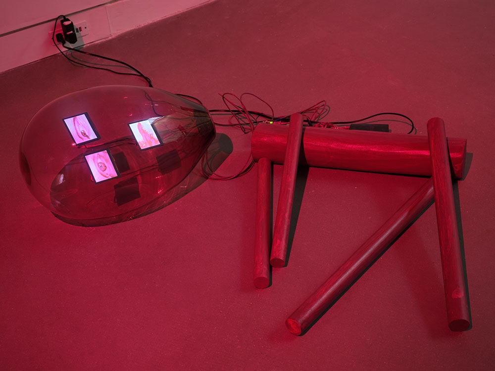 A red robot is collapsed on its side on the floor, dimly lit in red. The robot&rsquo;s body, appendages facing down to the right of the screen, is comprised of cylindrical tubes. To the left, its glass lightbulb-shaped head contains digital images of two eyes and a mouth. 