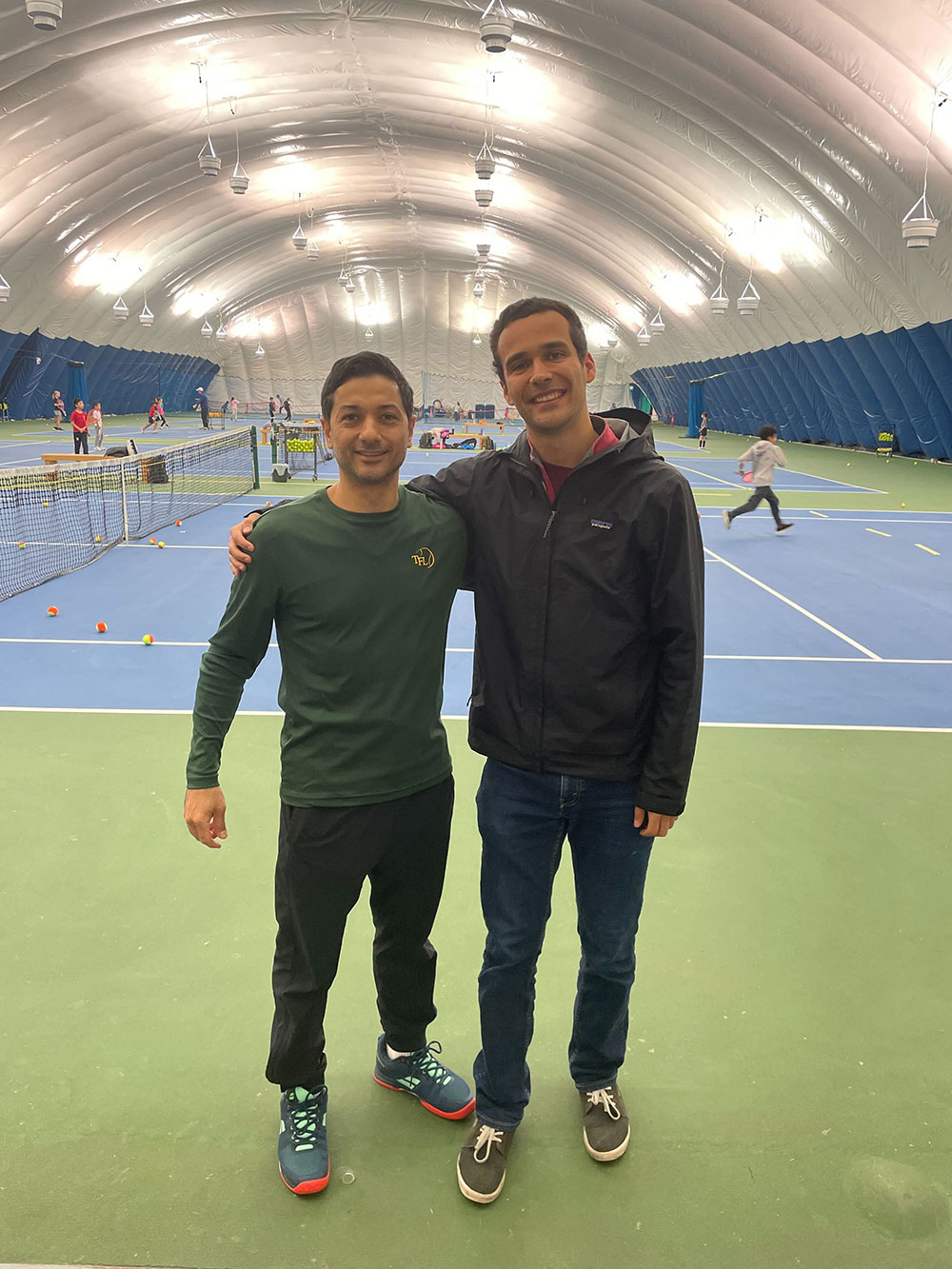 Hassan Askari, left, and Josh Kozelj, right stand with their arms around each other&rsquo;s shoulders. Hassan is wearing a dark green long-sleeved t-shirt. Kozelj is wearing a black rain jacket. They are both standing on green turf at the Coquitlam Tennis Centre, an indoor tennis centre with a domed white ceiling visible above them. Behind them, kids practice tennis on blue indoor courts with white lines.