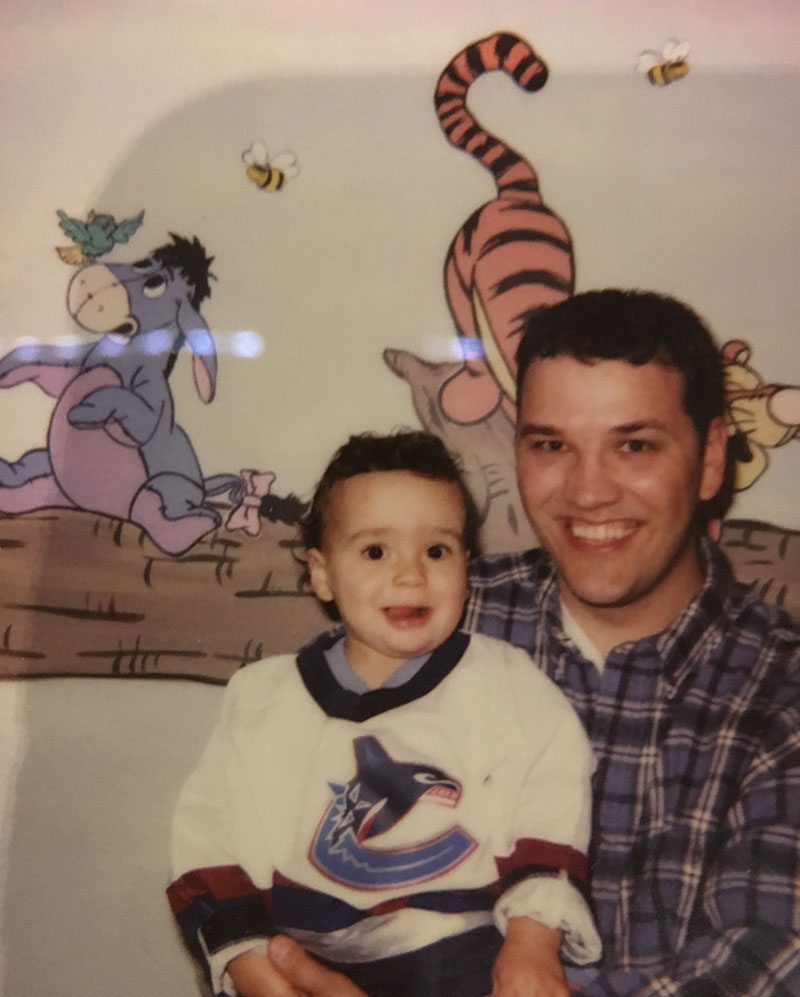 A family photo featuring Josh Kozelj, left, as a toddler. His father is holding him in his arms. Josh is wearing a white Vancouver Canucks hockey jersey. His father is wearing a blue plaid shirt. They are seated against a wall featuring a children&rsquo;s mural of characters from Winnie the Pooh.