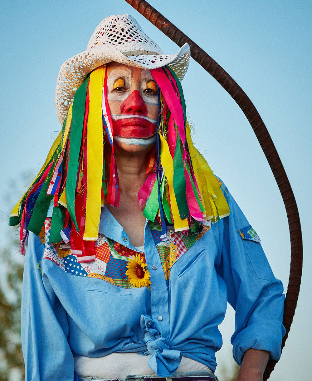 A woman looks directly at the camera. She is shot from the waist up. She is wearing a white straw hat, bright red, yellow and white clown makeup, and a wig made of long strips of yellow, fuchsia, blue and red fabric. She is wearing a light blue cowboy shirt with a rainbow patchwork quilt pattern across her chest.