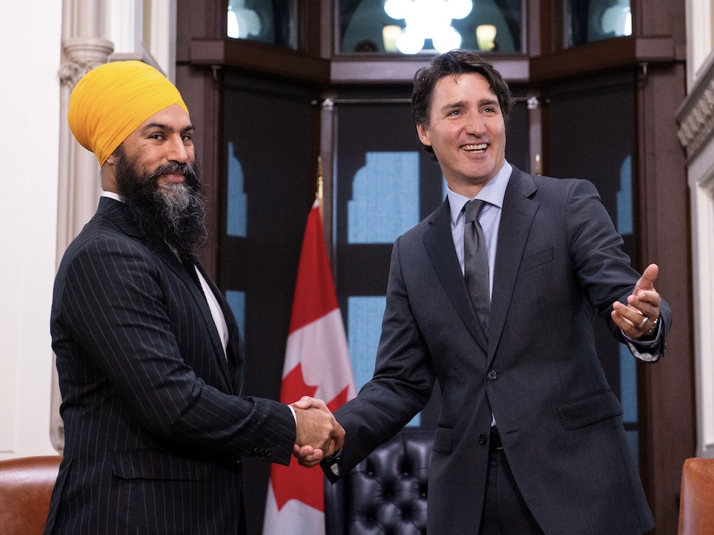 Jagmeet Singh shake hands with Justin Trudeau. They are standing together in front of a Canadian flag.