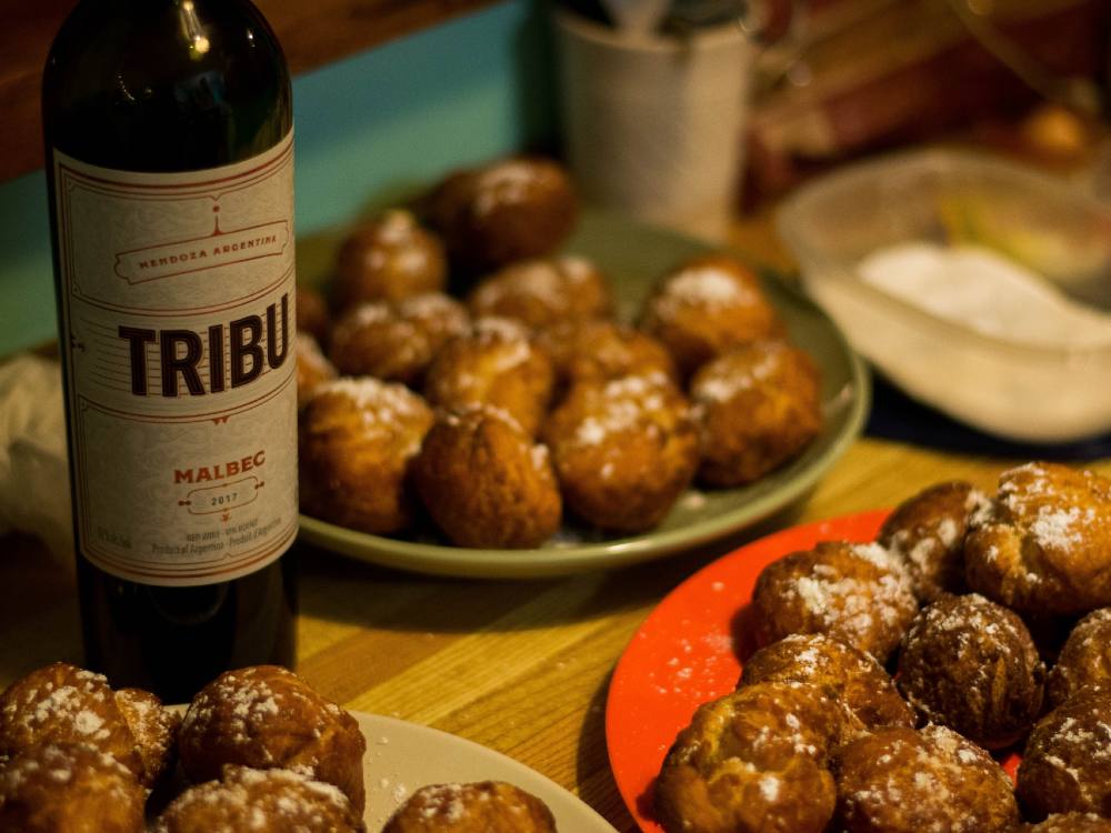 An artsy photo of a bottle of malbec on a table, surrounded by homemade doughnut holes dusted in icing sugar.
