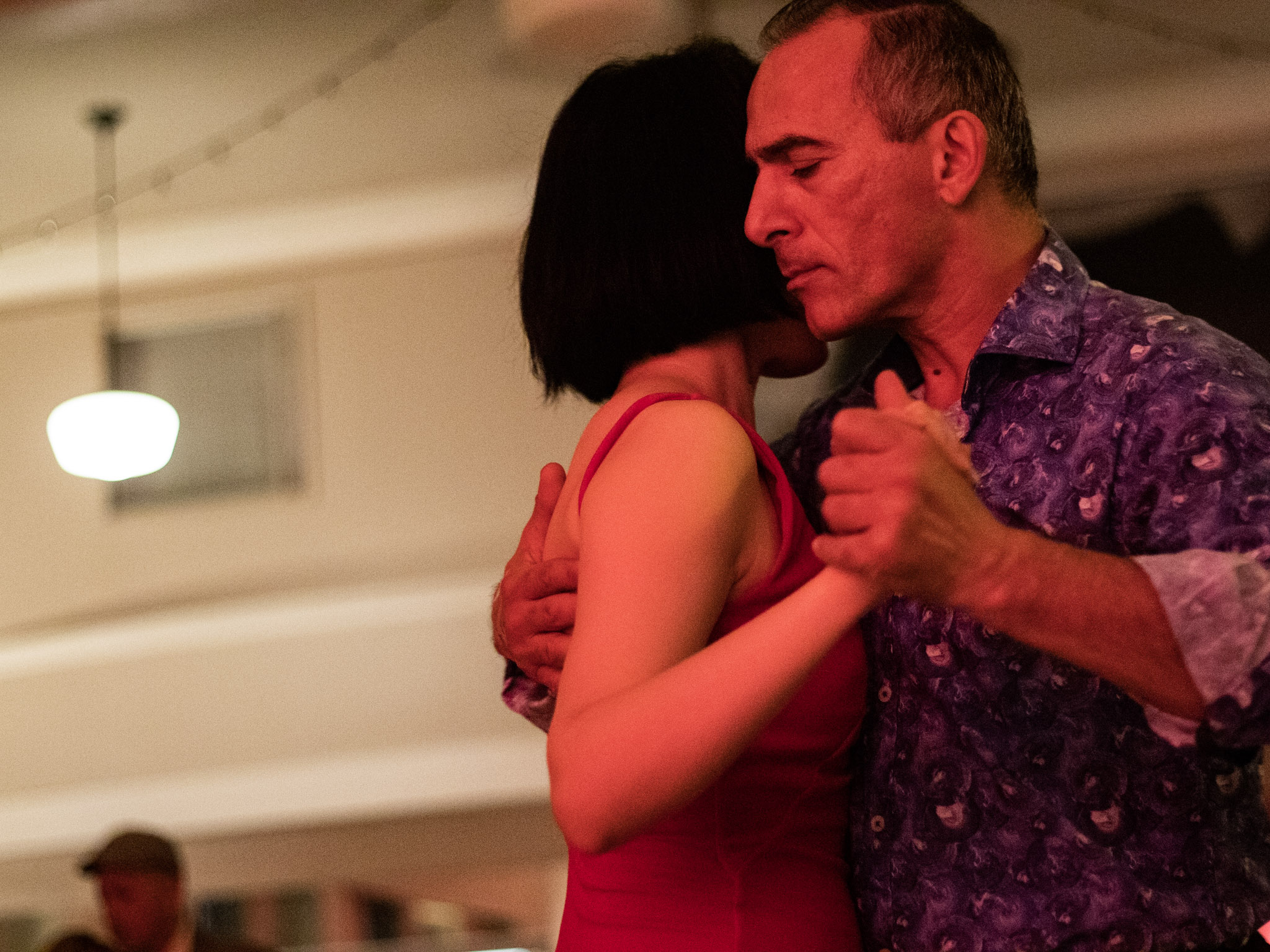 ID: A close-up photograph of a man and a woman dancing. The woman’s face is hidden behind the man’s face, his eyes are closed as their chests are pressed together and their hands clasped.