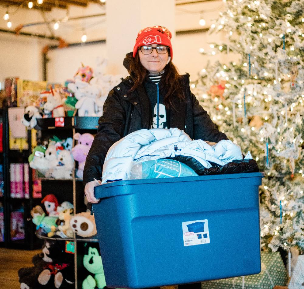 A woman wearing glasses and a red toque examines and item with a red ribbon amidst shelves of other items.