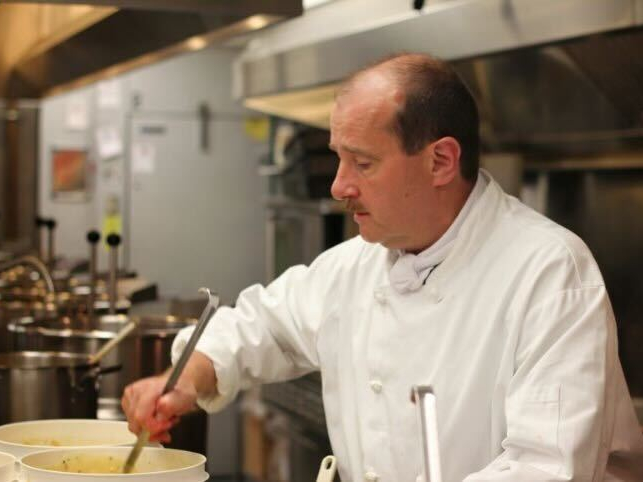 A man in chef’s whites stirs an industrial white bucket of soup with a long stainless steel ladel. In the background is a hood range fan and many large stainless steel stockpots.