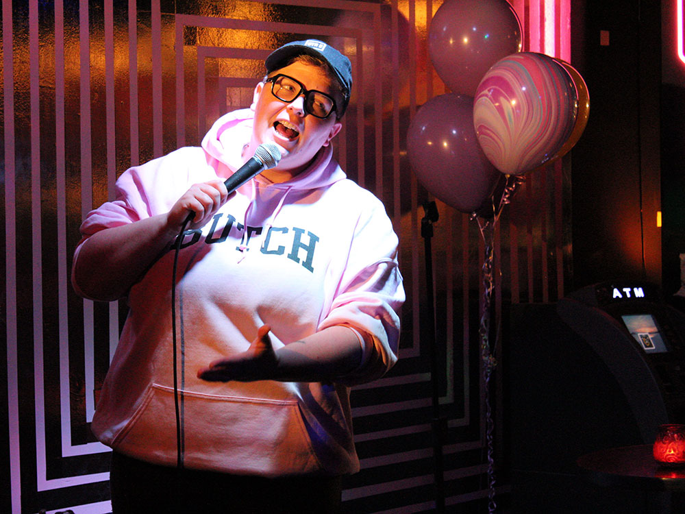 Jackie Hoffart wears a light pink hoodie with the word “Butch” in varsity script across the chest. They are wearing a ball cap and speaking into a microphone. Behind them is a dim wall and a bunch of balloons.