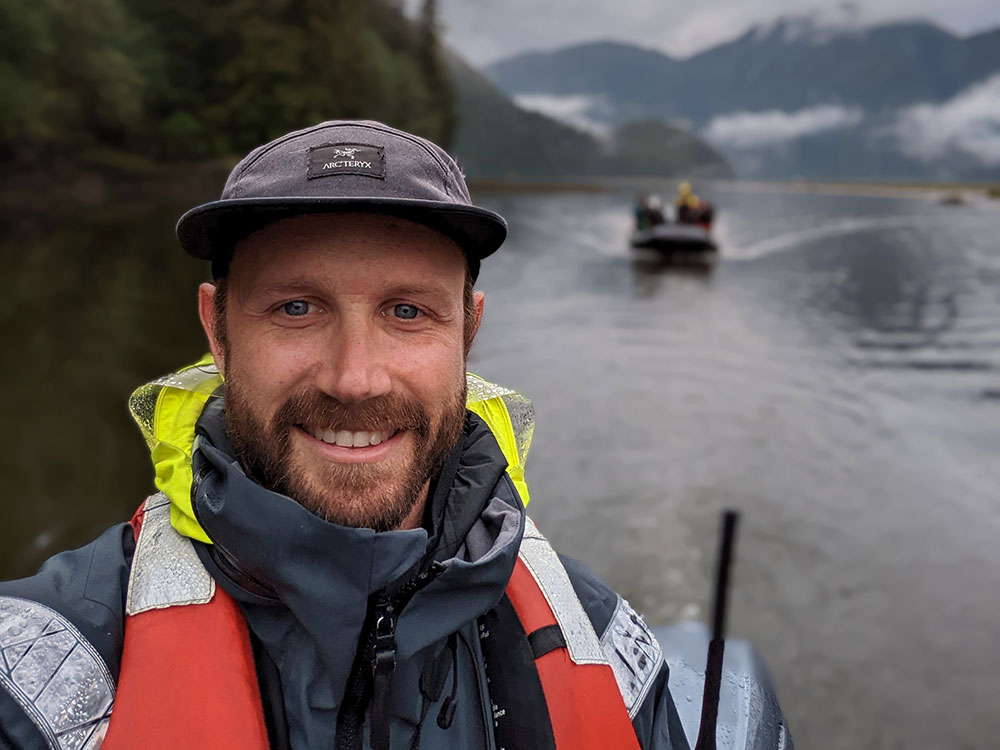 A person wearing a ball cap, rain gear and a life jacket, holding a walkie talkie, smiles at the camera. Behind him, a dinghy full of people is visible but blurry in the background.
