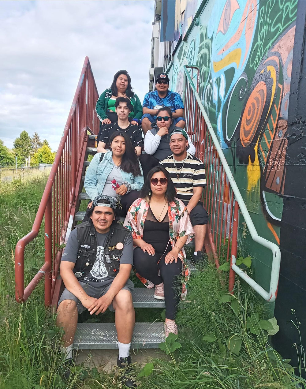 Alex Taylor-McCallum is seated on the bottom stair, to the left of the staircase, with seven people next to one of his pieces of artwork. Long green grass grows around the stairs. It&rsquo;s a sunny day and people are looking at the camera, smiling.