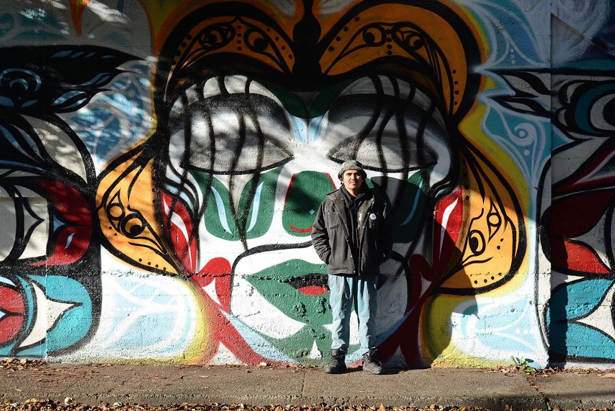 Alex Taylor-McCallum stands in the sun in front of one of his murals in Esquimalt, a stylized illustration of a person&rsquo;s face. The illustration uses black, orange, white, green and red spray paint. McCallum stands with his hands in the pockets of his black denim jacket. He is wearing a hat and blue jeans.