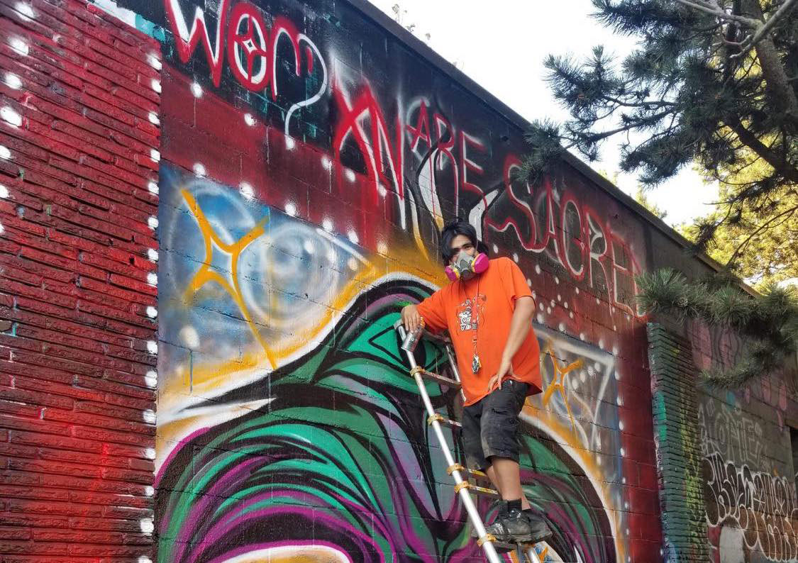 Alex Taylor-McCallum in an orange T-shirt and a respirator at the top of a ladder against a colourful graffiti wall. He is looking down towards the camera and holding a can of spray paint. The wall reads &ldquo;Womxn are sacred&rdquo; in handwritten white and red script across the top, below the text is a stylized purple and green illustration of a hummingbird. 