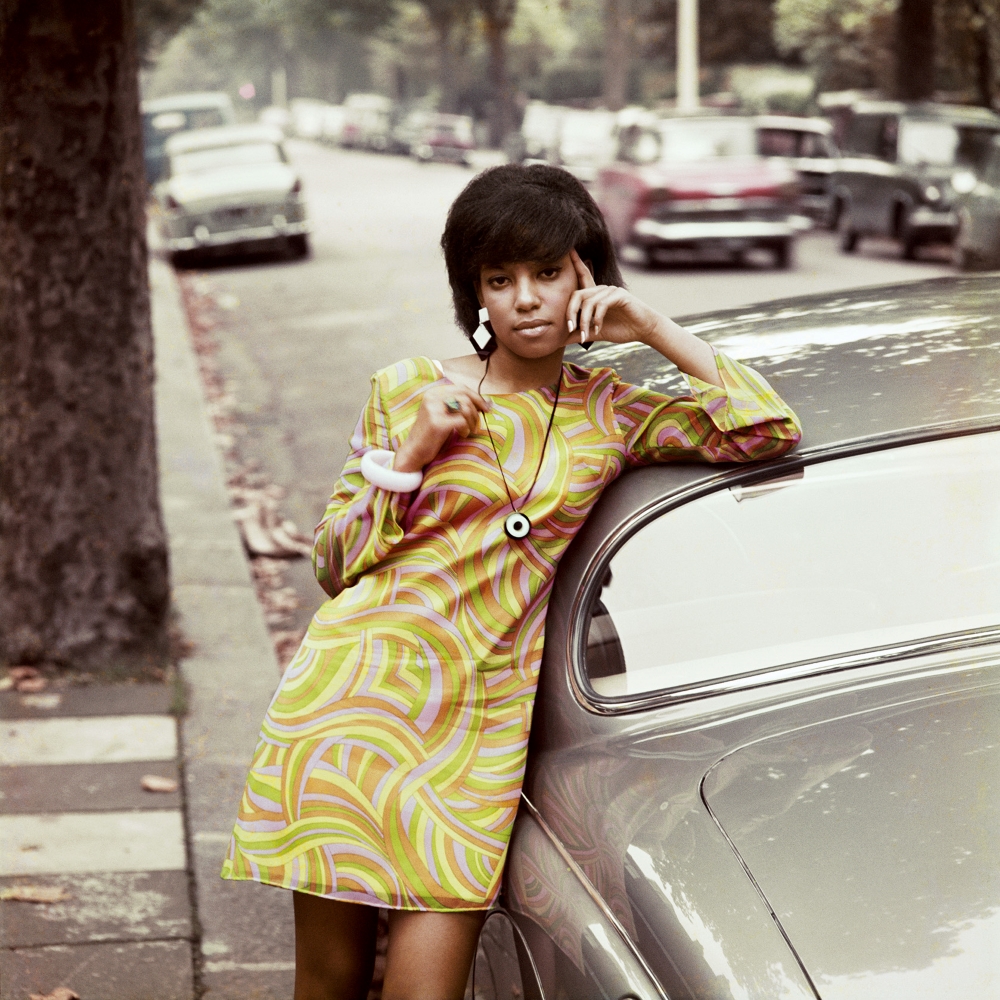 A young Black woman in a long-sleeved dress in a swirling orange, yellow and green &lsquo;60s motif leans against a grey car on a quiet residential street. 
