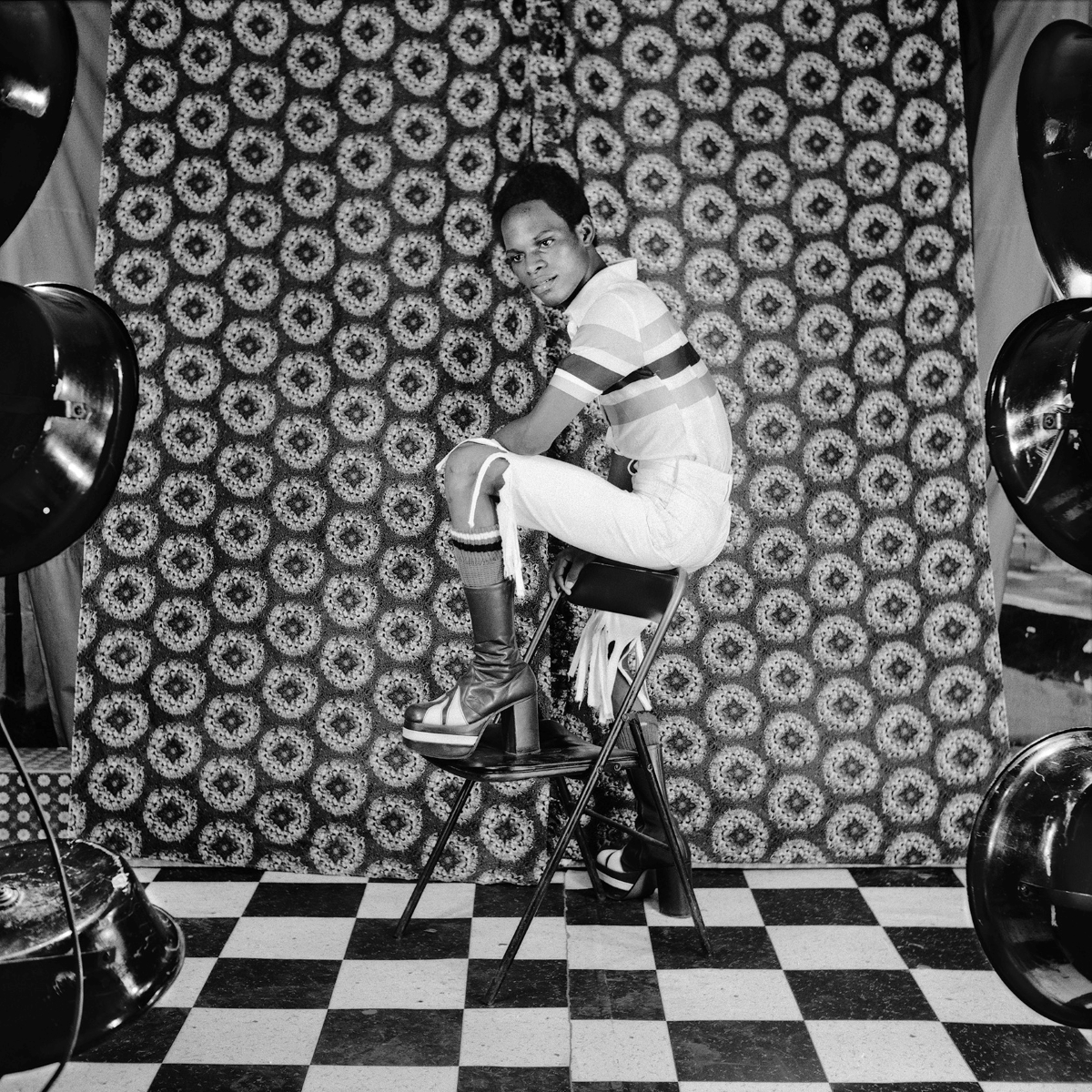 A black and white portrait of a young Black person with short hair, a polo T-shirt, disco boots and fringe shorts posing on a plastic chair. They are standing against a &lsquo;70s patterned wallpaper with circular designs.
