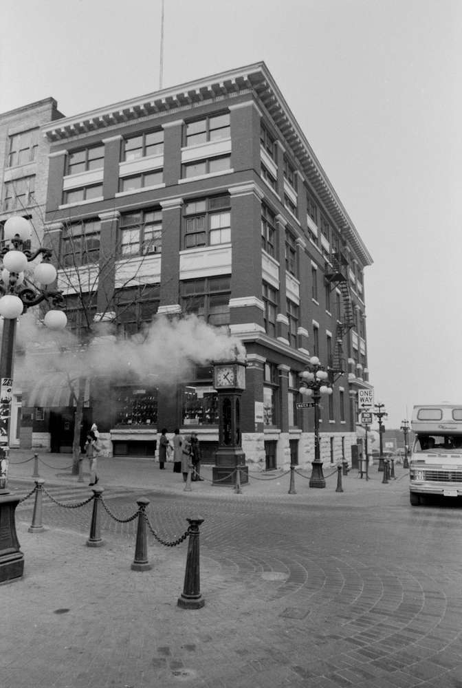 A black-and-white photo shows people standing around the steam clock as it vents steam.
