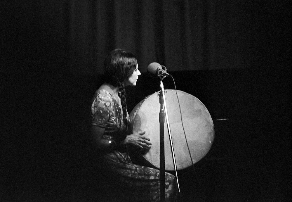 A black and white photo of Alanis Obomsawin, an Indigenous woman with long braided hair, sitting onstage and holding a drum in front of a microphone. She is facing the right of the frame.