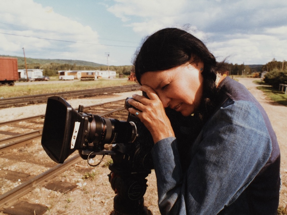 An Indigenous woman with long dark braided hair leans forward toward the left of the screen to look into a camera. She is outdoors on a sunny day near a railroad track, wearing a denim shirt.