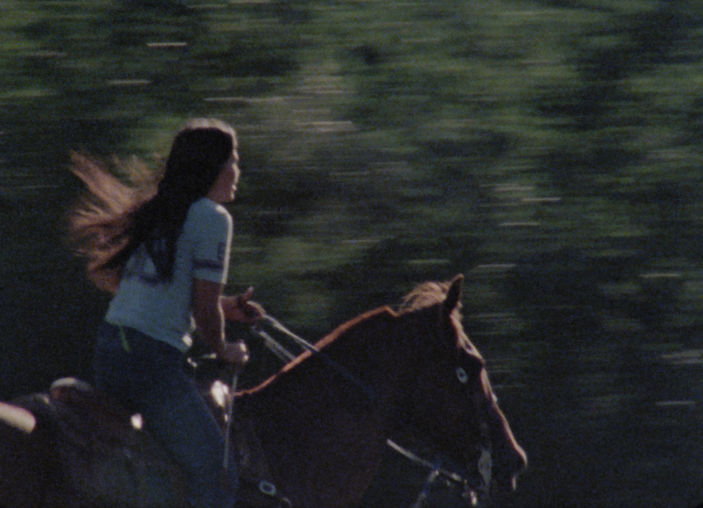 An Indigenous woman on horseback moves towards the right side of the screen. Her long hair blows behind her and she is riding a brown horse. The background is green and blurry due to the motion captured on film.