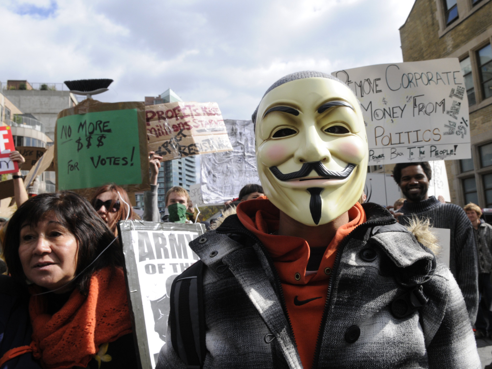 A person wearing a Guy Fawkes mask looks at the camera amid a protest on corporate investment in politics. Signs read 'Remove corporate money from politics&rdquo; and &ldquo;No more $$$ for votes!'