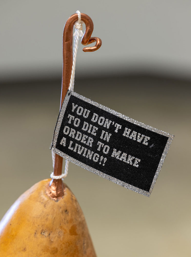 The vertical bronzed stem at the top of a pear-shaped golden gord holds a black flag with silver text in capital letters that reads, 'You don&rsquo;t have to die in order to make a living!!'