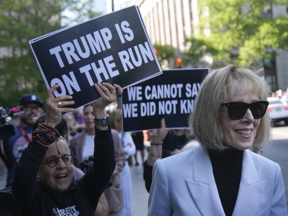 A woman with short blonde hair, sunglasses and a light grey blazer over a black turtleneck walks down a New York City sidewalk, smiling. Behind her, demonstrators hold black placards with white sans-serf typeface that read &ldquo;Trump is on the run&rdquo; and &ldquo;We cannot say we did not know.&rdquo;