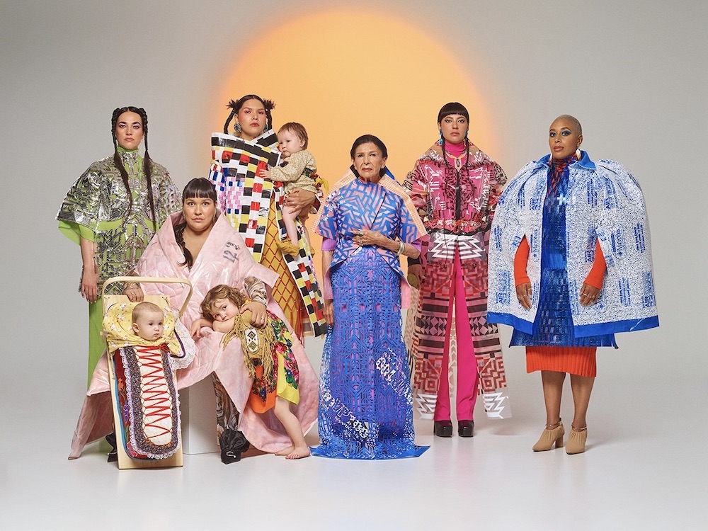A group of Black and Indigenous women and children stand together wearing colourful, futuristic clothing in a studio with white floors and a grey background. To the left, a baby is bundled in a futuristic wooden sled.