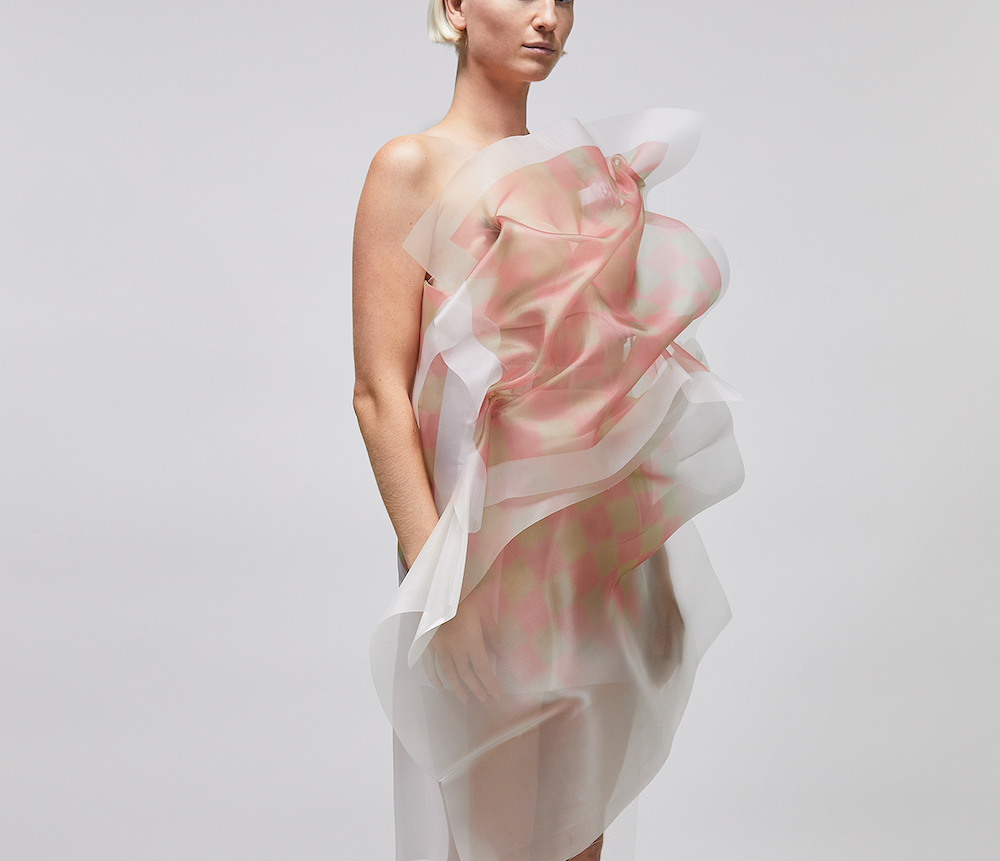 A woman with short blonde hair models an off-the-shoulder dress made of transparent, billowy fabric. We can&rsquo;t see her eyes, and she stands against a light grey background.