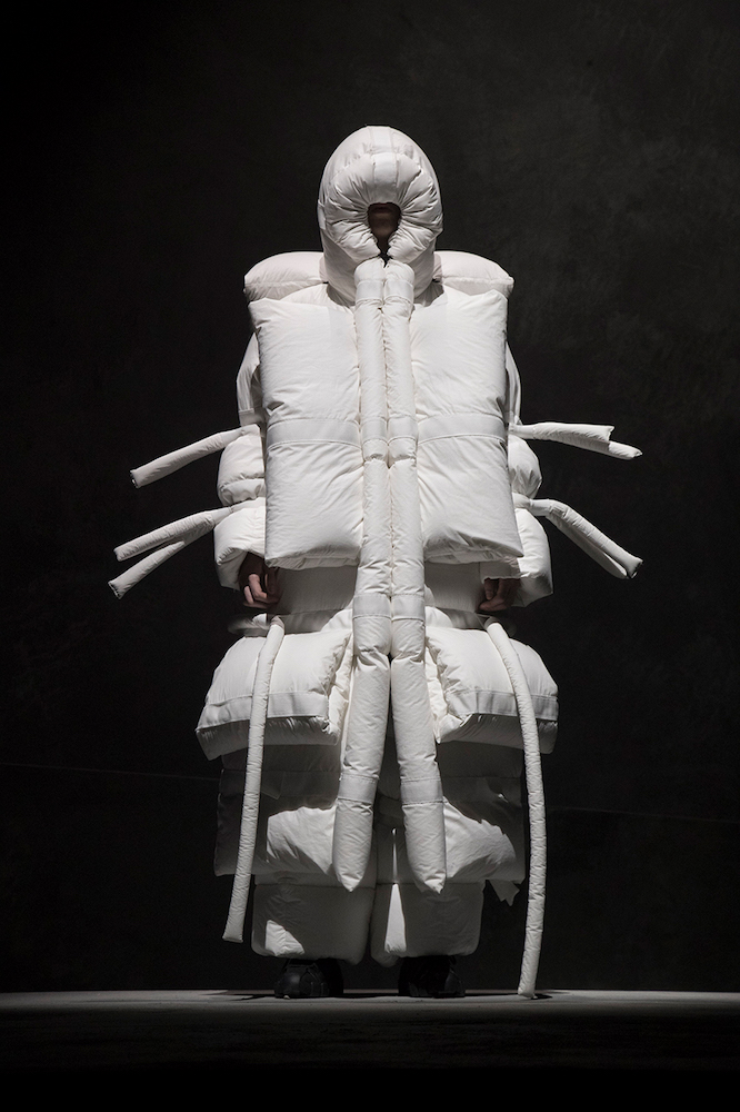 A model stands against a dark grey background in a full-body oversized puffy white jacket with white tubes extending from the elbows down the sides of the body. Their face is barely discernible inside the recesses of a deep hood.