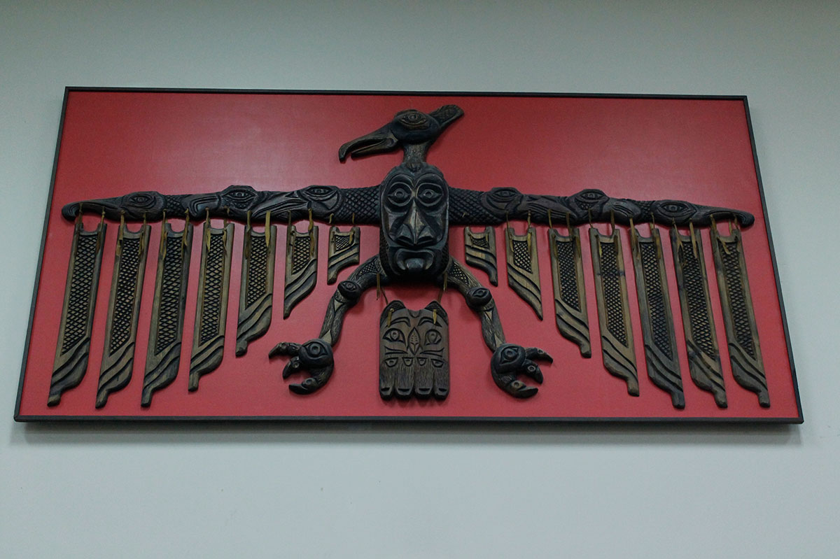 An elaborate wooden bird carving is mounted on a red background, and hung on a white wall.