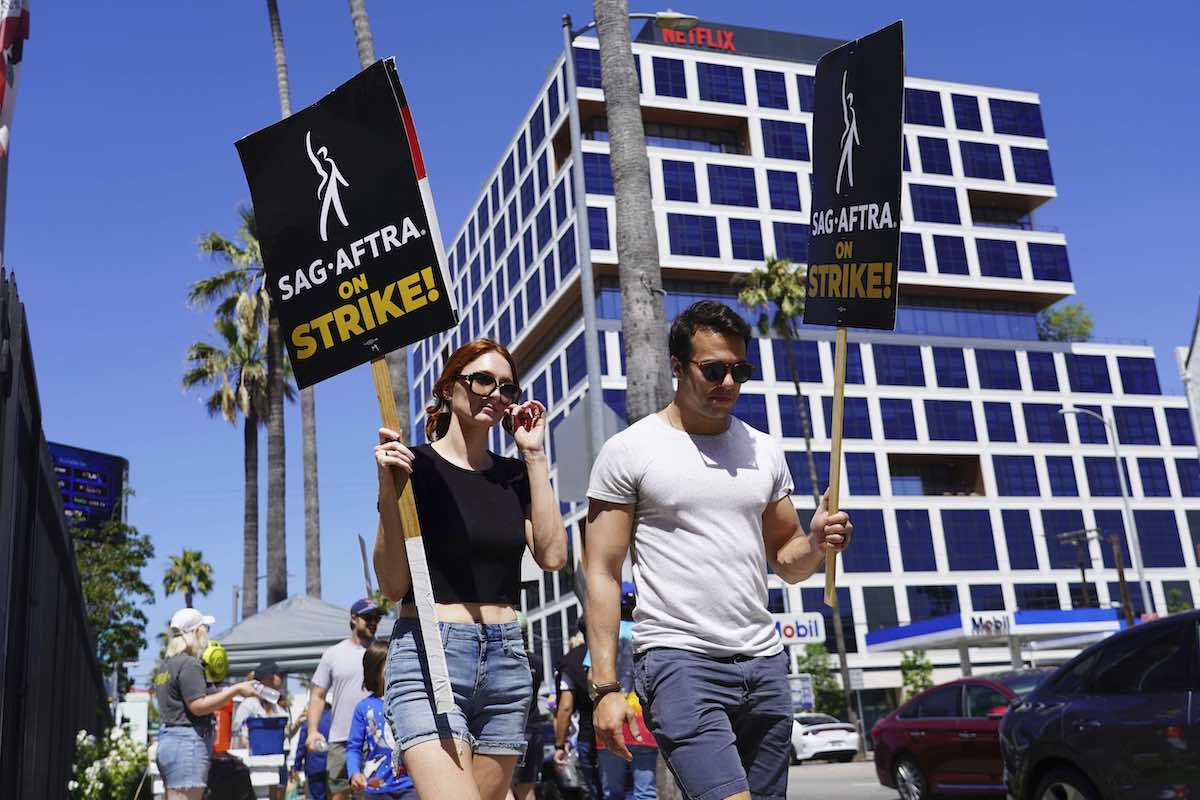 On the left, a woman with red hair, sunglasses, a black T-shirt and jean shorts holds a black picket sign that reads &ldquo;SAG-AFTRA on strike!&rdquo; She is walking beside a dark-haired man in sunglasses and a white T-shirt and grey shorts holding a similar sign. They are walking on the sidewalk in front of a large office building with square blue windows and cube-shaped white borders on a bright sunny day.