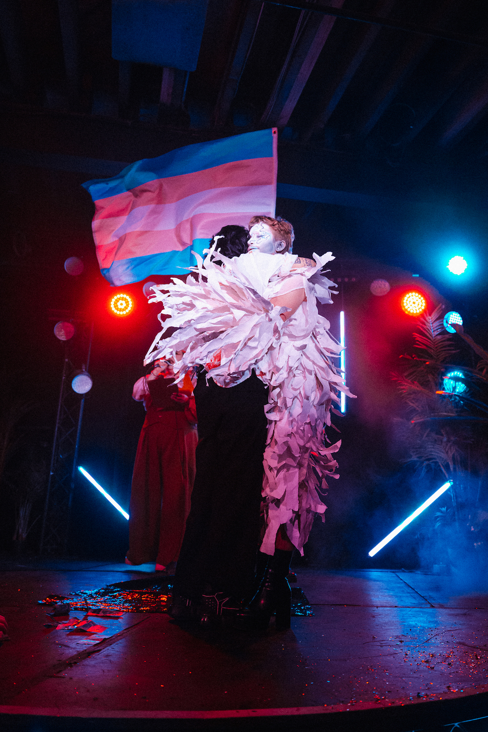 The signature trans flag, with its white, pink and blue colours, sways underneath the lights as two drag performers hug on stage.