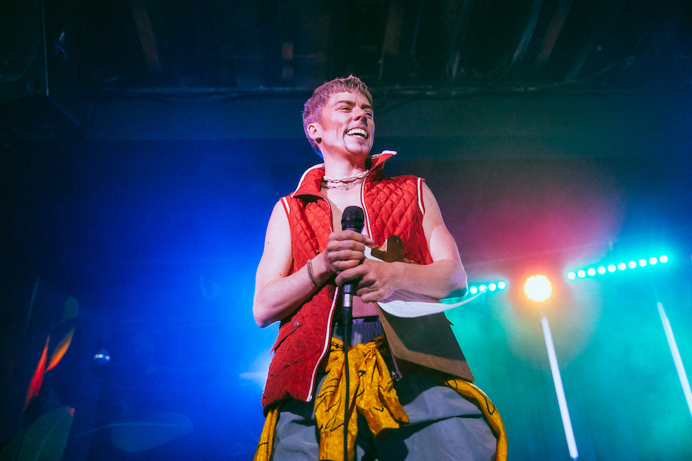 A person in an orange sleeveless jacket is lit by stage lights. They are holding a microphone and a clipboard. They are smiling widely.