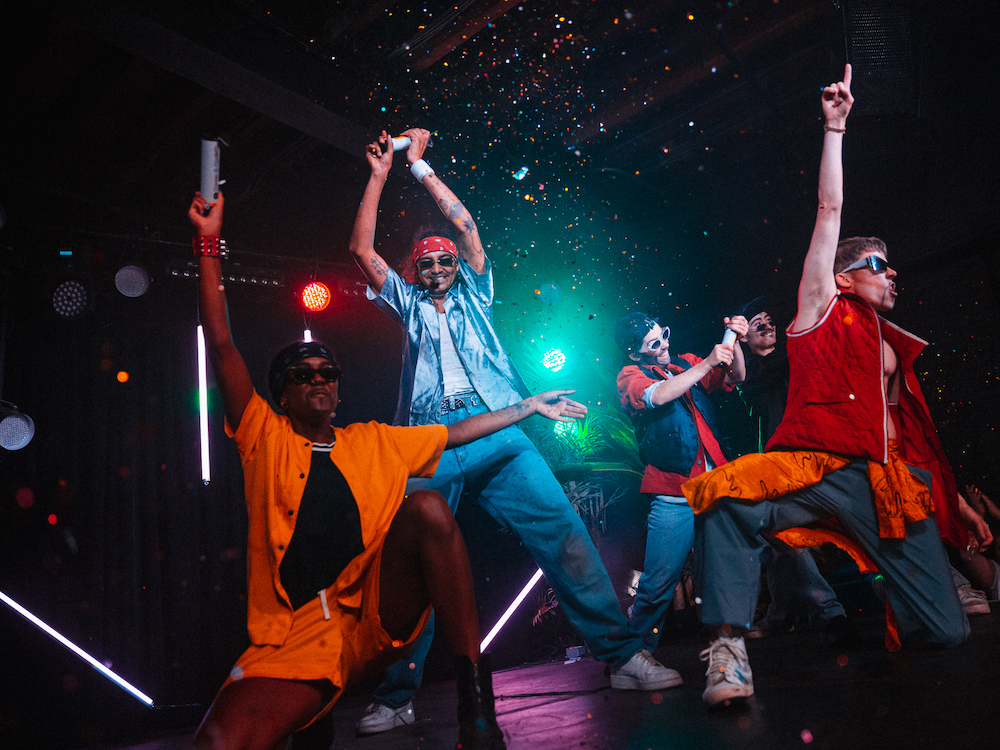 A group of smiling performers, dressed as a boy band, dance in the glow of a turquoise stage light as confetti fills the air.