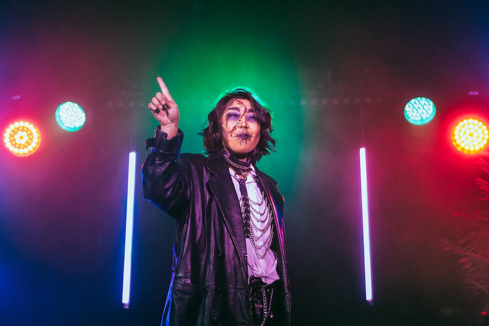A performer in a black leather jacket and adorned in chains wags their finger. Their makeup has created the visual effect of a bloody gash across their face and purple sunken eye sockets.