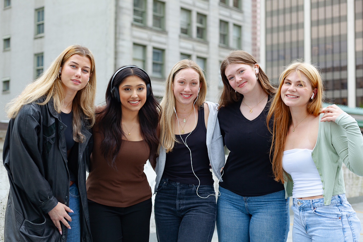 Five young women wearing headphones stand together with their arms around each other&rsquo;s shoulders.