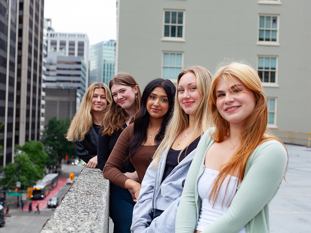 Five young women stand shoulder to shoulder against a concrete barrier that comes up to their waists, looking right towards the camera.