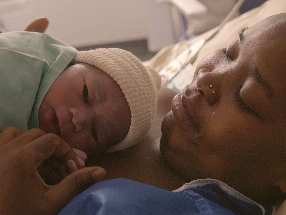 A closeup of a woman and her newborn. The newborn&rsquo;s hand is holding their parent&rsquo;s finger.