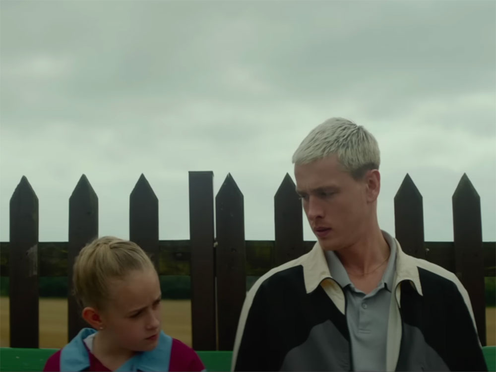 A girl in a blond French braid and a light blue and maroon soccer jersey sits to the left of the frame next to a young man in a cream, grey and black jacket over a grey polo shirt. He has short bleached blond hair. They are sitting outside against a dark picket fence. In the background, the sky is grey.