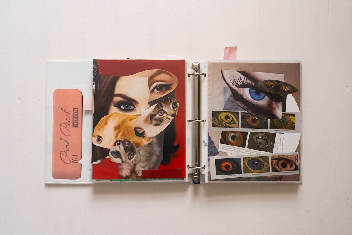 A large white binder lies open against a white background. To the left, an oversized Pink Pearl eraser is mounted onto the side of the binder. The binder features two pieces of collage work under thin plastic sheaths. They feature colour photo cut-outs of human and animal eyes.