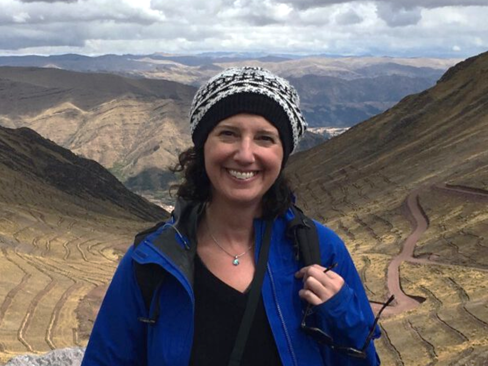 A woman in a toque and blue jacket smiles in the foreground, with brown hills and clouds in the background.