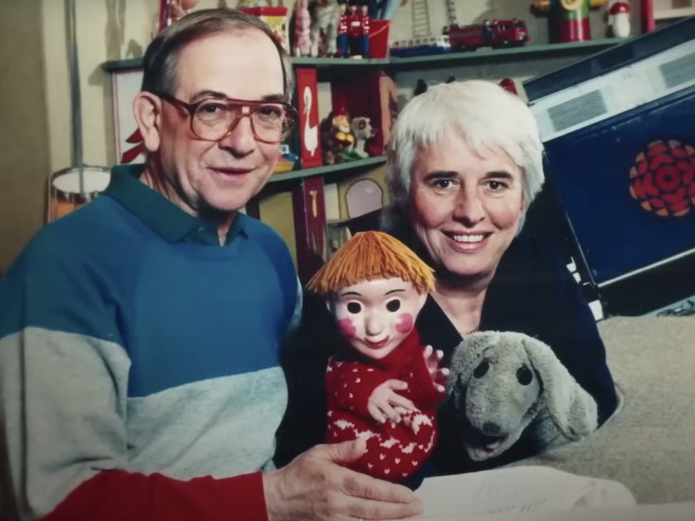 An archival photo of two adults on a television sound stage. On the left, Ernie Coombs has short dirty blond hair and red glasses. The collar of his green polo shirt is visible under his blue, grey and red striped pullover. On the right, Judith Lawrence has short white hair and is looking directly at the camera, smiling. She is wearing a black top and holds two puppets with Coombs: a child with red hair, and a grey dog. 
