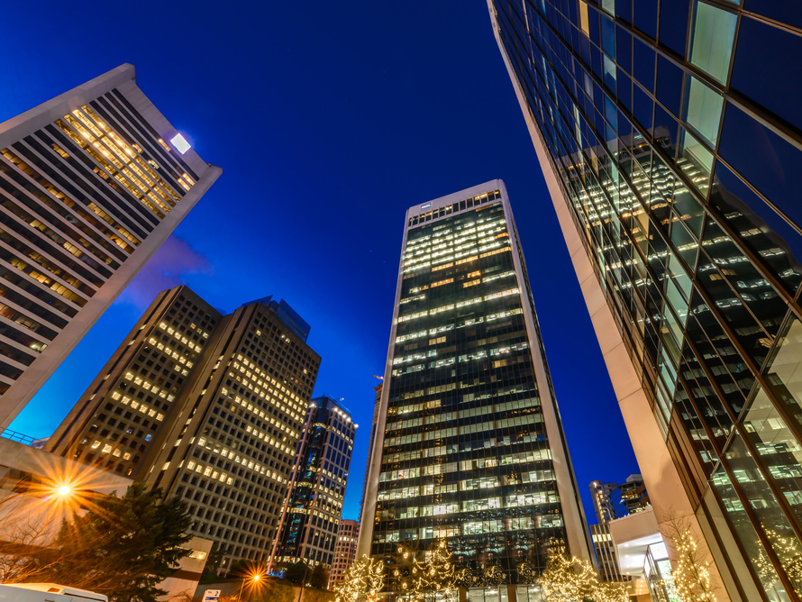 At night, looking up at tall glass office buildings with window lights glowing yellow.