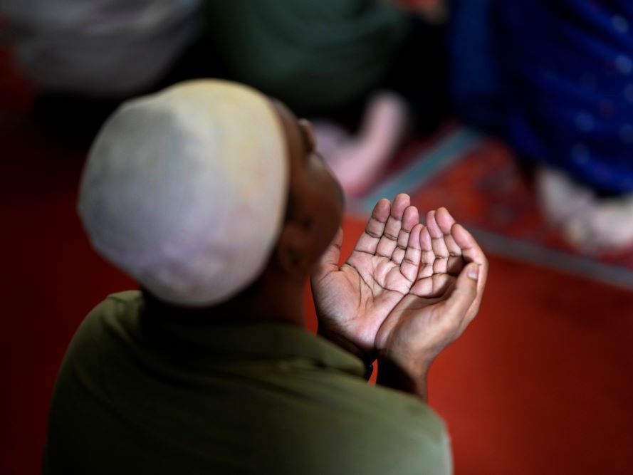 A man wearing a white prayer cap and a green shirt prays with his hands held in front of him, palms up.