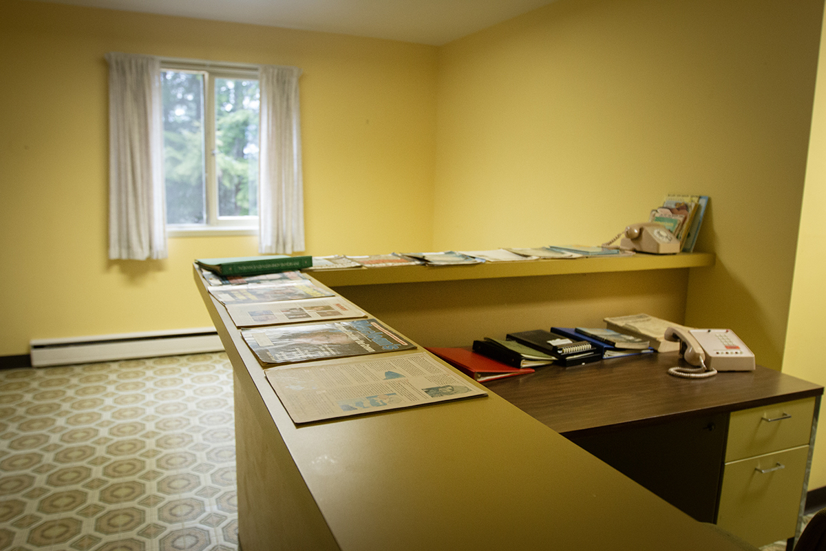 A room with yellow walls and yellow patterned linoleum floors. There is a counter with two telephones, one of them rotary dial, and magazines.