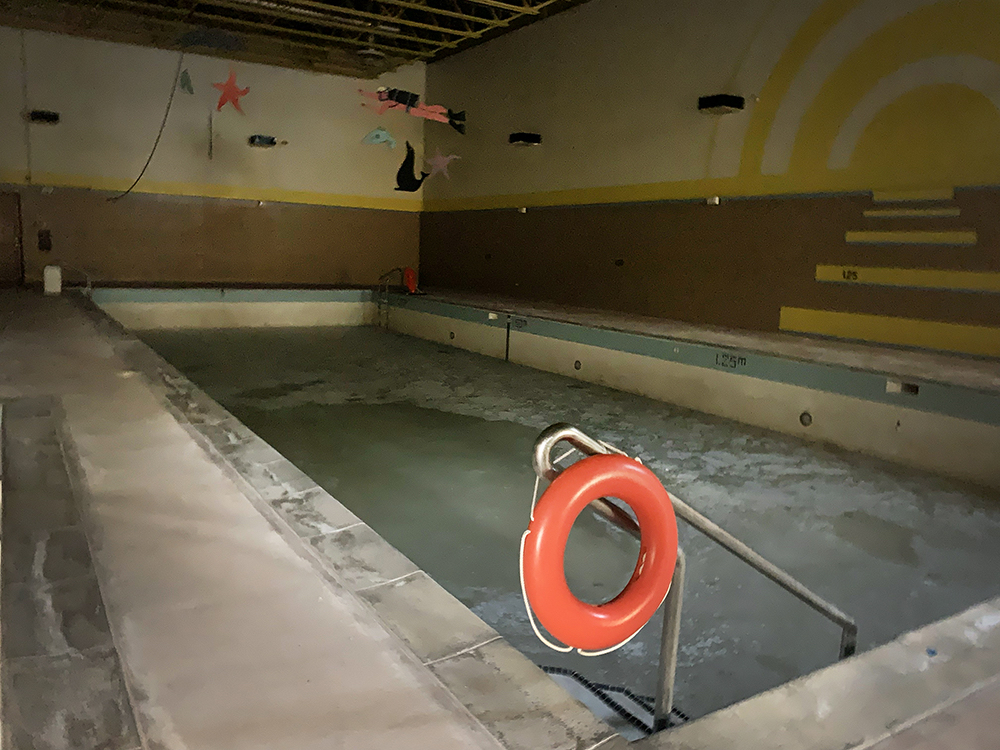 An empty swimming pool sits in a darkened building. There is a red life-saving buoy hanging from the pool&rsquo;s hand rail.