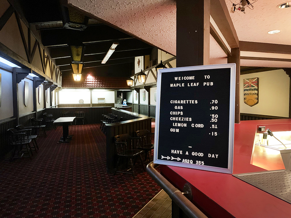 A darkened room with tables, chairs and a dartboard. A sign says, &ldquo;Welcome to Maple Leaf Pub.&rdquo; 