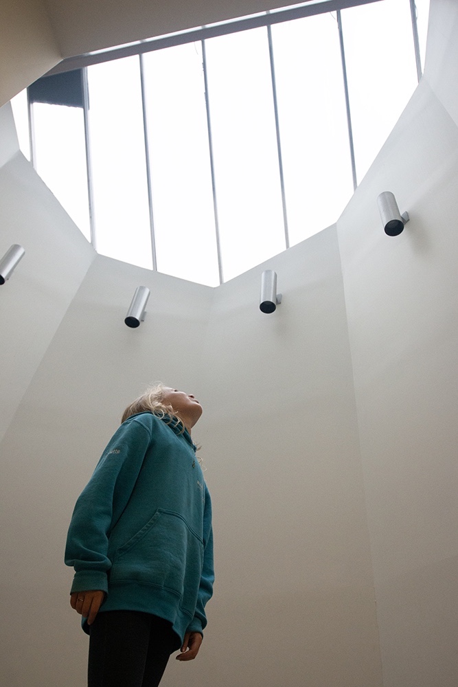 A girl stands beneath a towering glass ceiling looking upward.